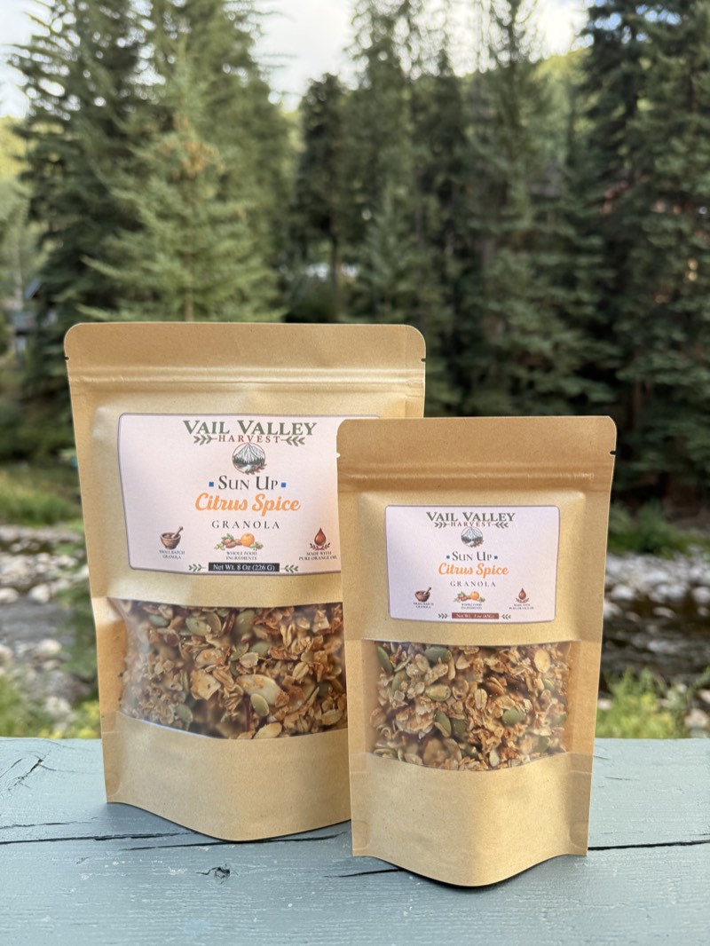 Sun Up Citrus Spice granola pouch in front of Vail pines