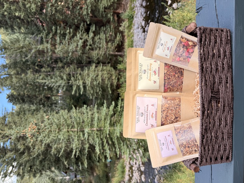 A wicker gift basket holding four Vail Valley Harvest granola pouches, with Gore Creek behind