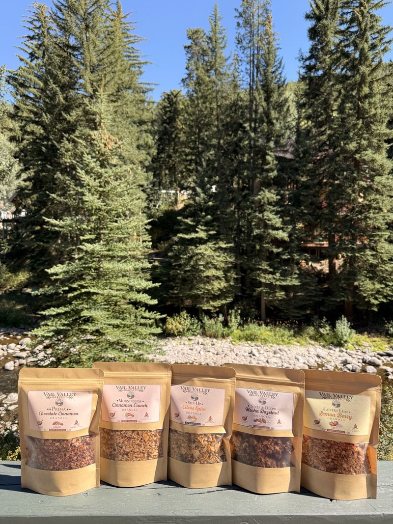 The five Vail Valley Harvest granola pouches lined up on a deck rail against Vail pines